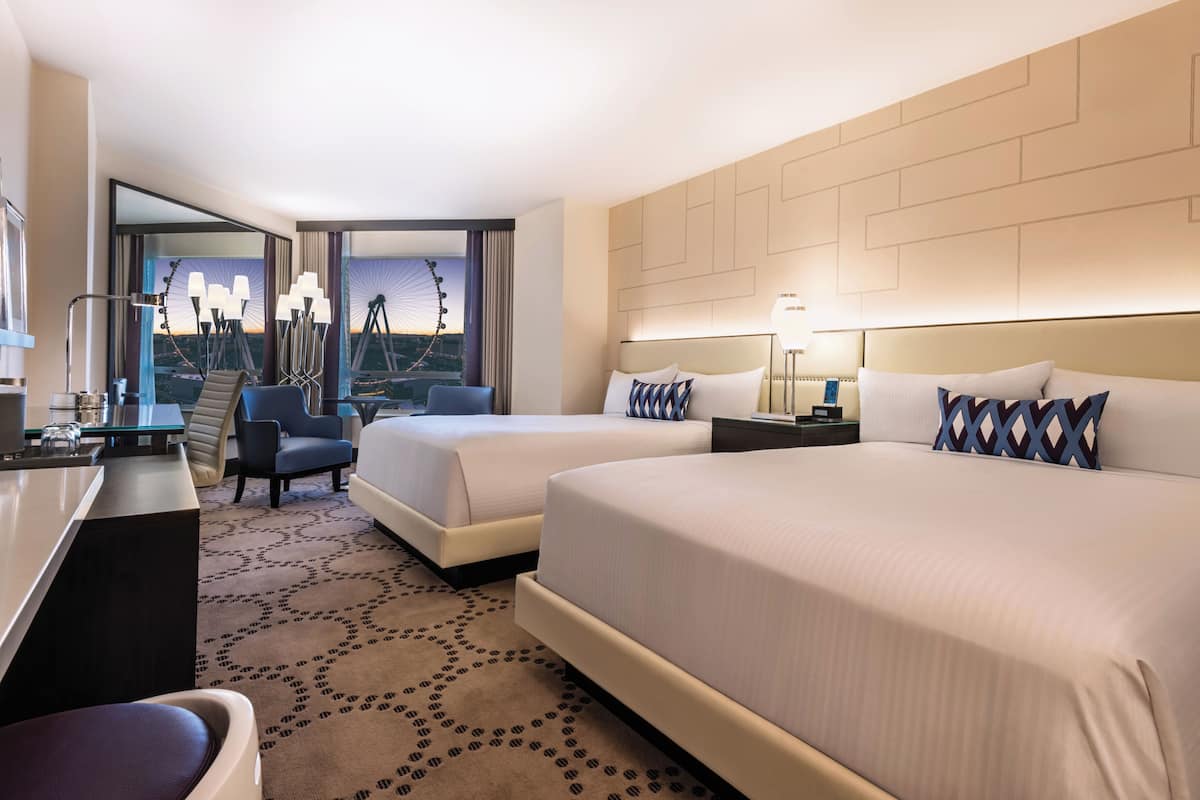 A hotel room with two beds, a desk, and a chair. There is a view of a sunset through a window.