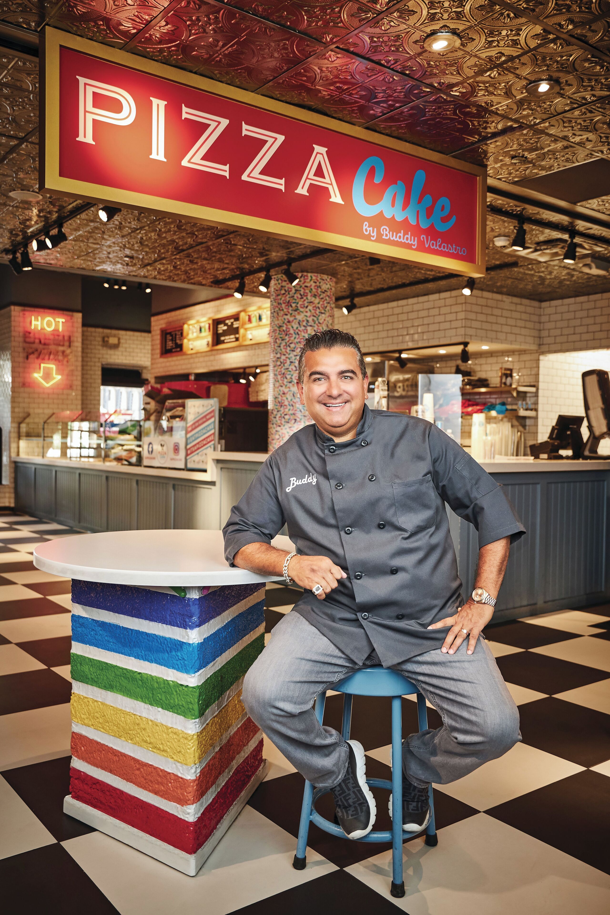 A chef seated at a colorful, striped stool in a restaurant with a 'PIZZA Cake' sign above.