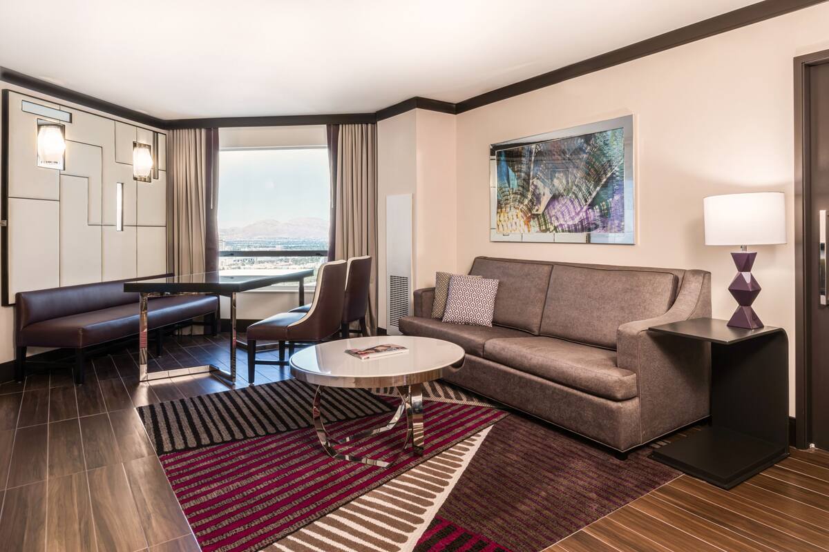 A modern hotel room with a sofa, a small table, a coffee table, and a bed. There is a painting on the wall and a window with a view of the city.