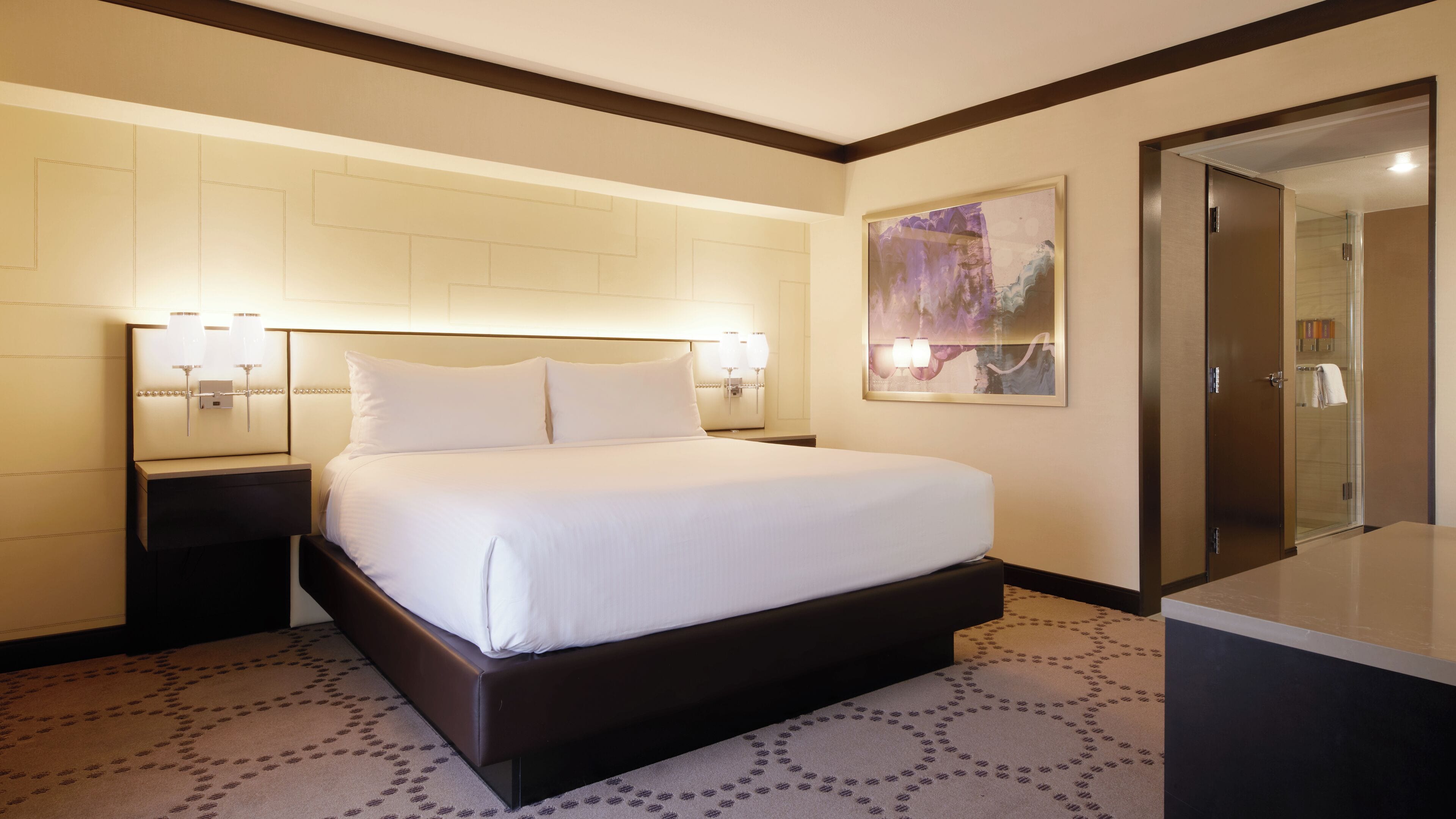A hotel room with a large bed, two bedside tables, a wall-mounted light fixture, a painting, and a mirror.