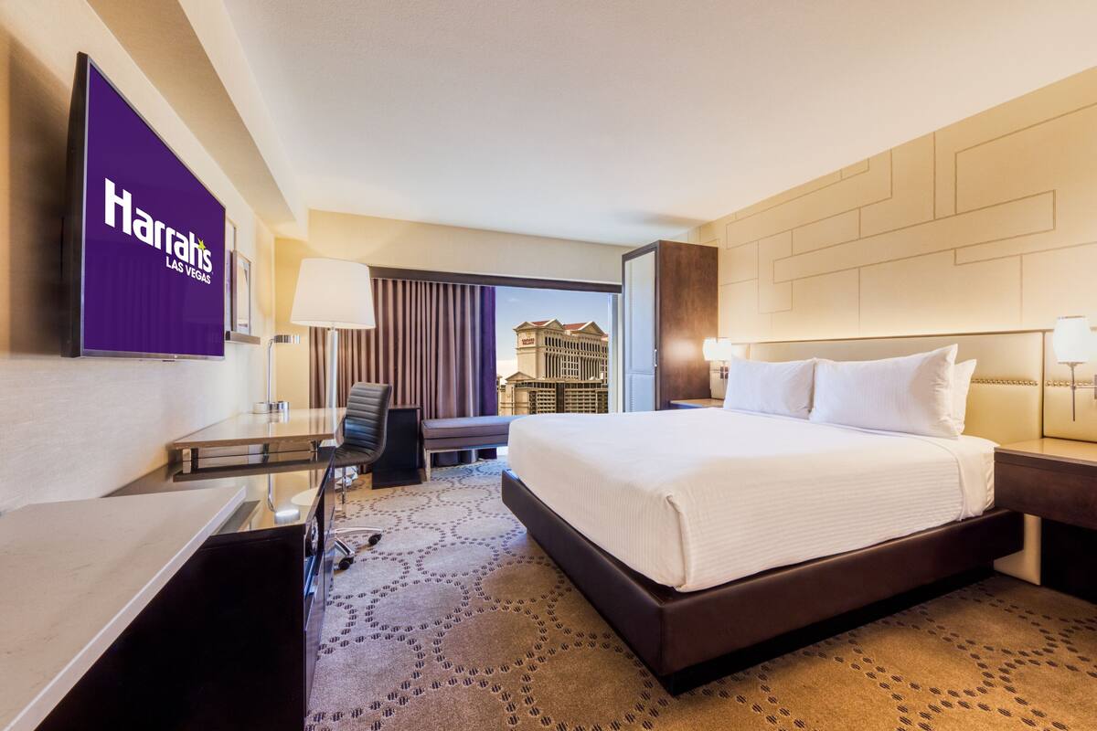 A hotel room with a large bed, a desk, a chair, a lamp, and a view of a building through the window.
