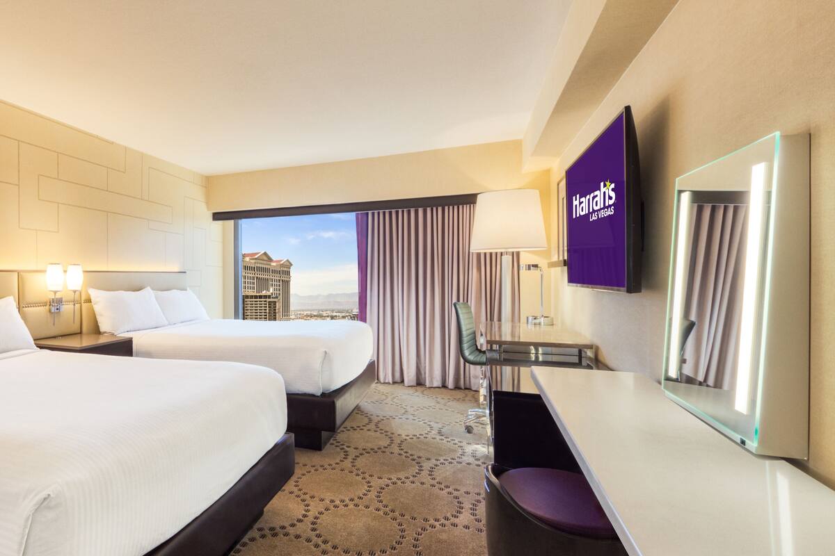 A hotel room with two beds, a desk, a chair, a TV, and a view of a cityscape.