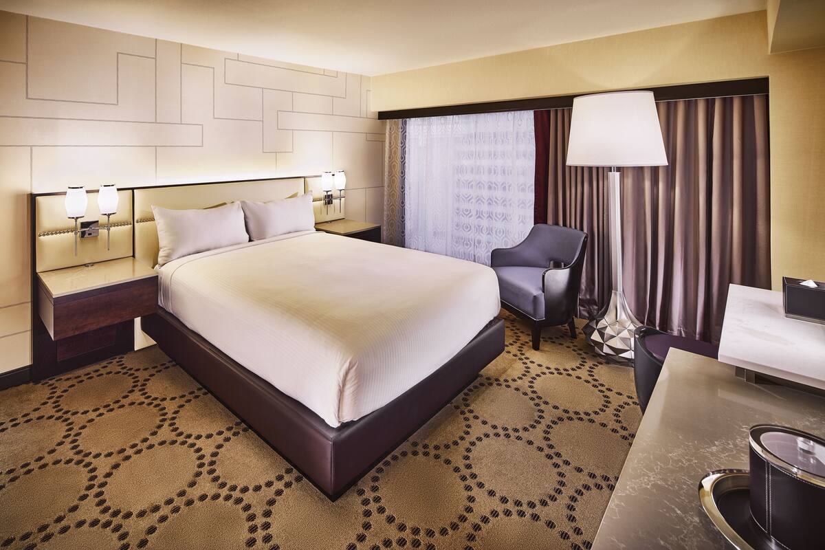 A hotel room with a large bed, a desk, a chair, a lamp, and a patterned carpet.