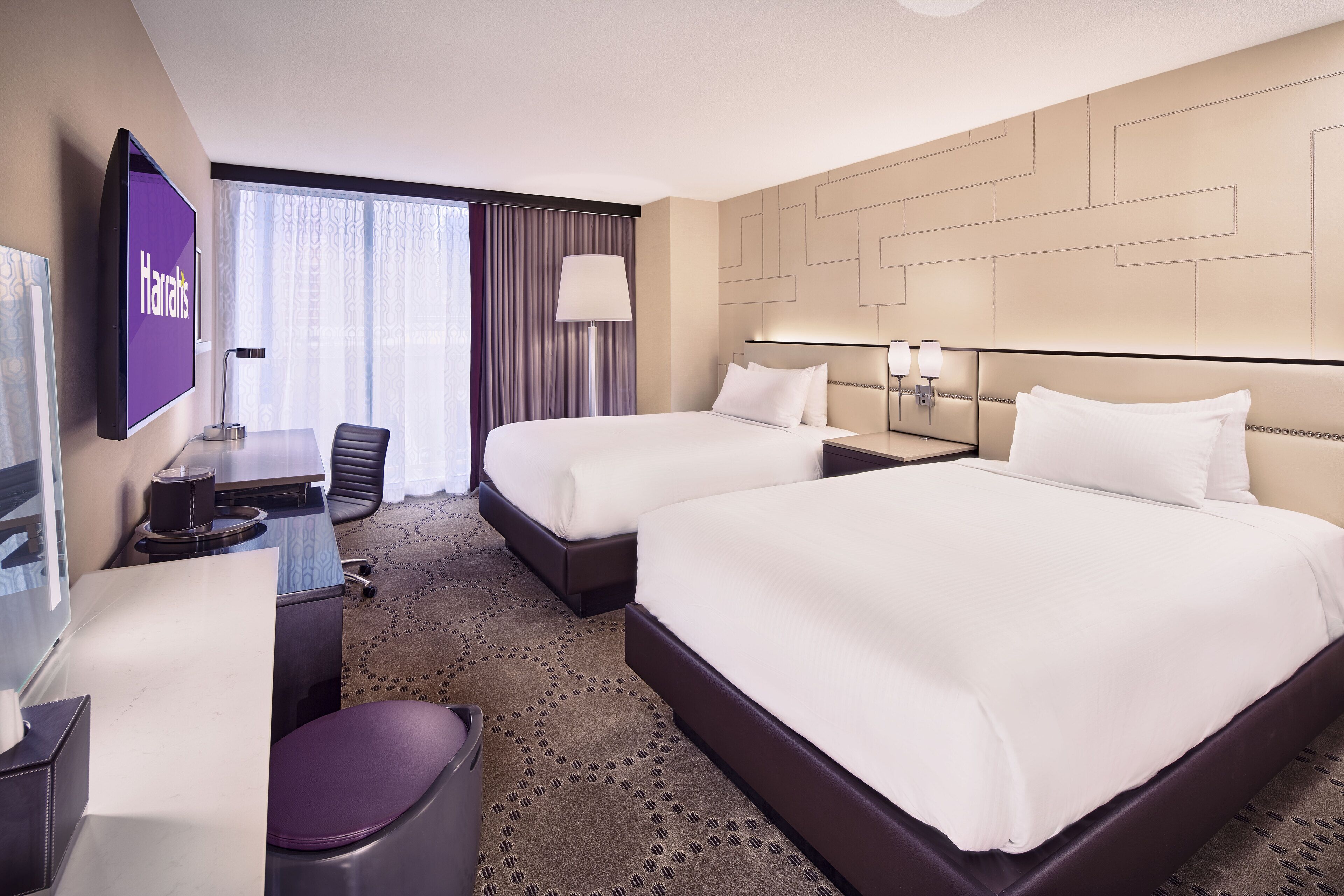 A hotel room with two beds, a desk, a chair, a TV, and a large window with curtains.