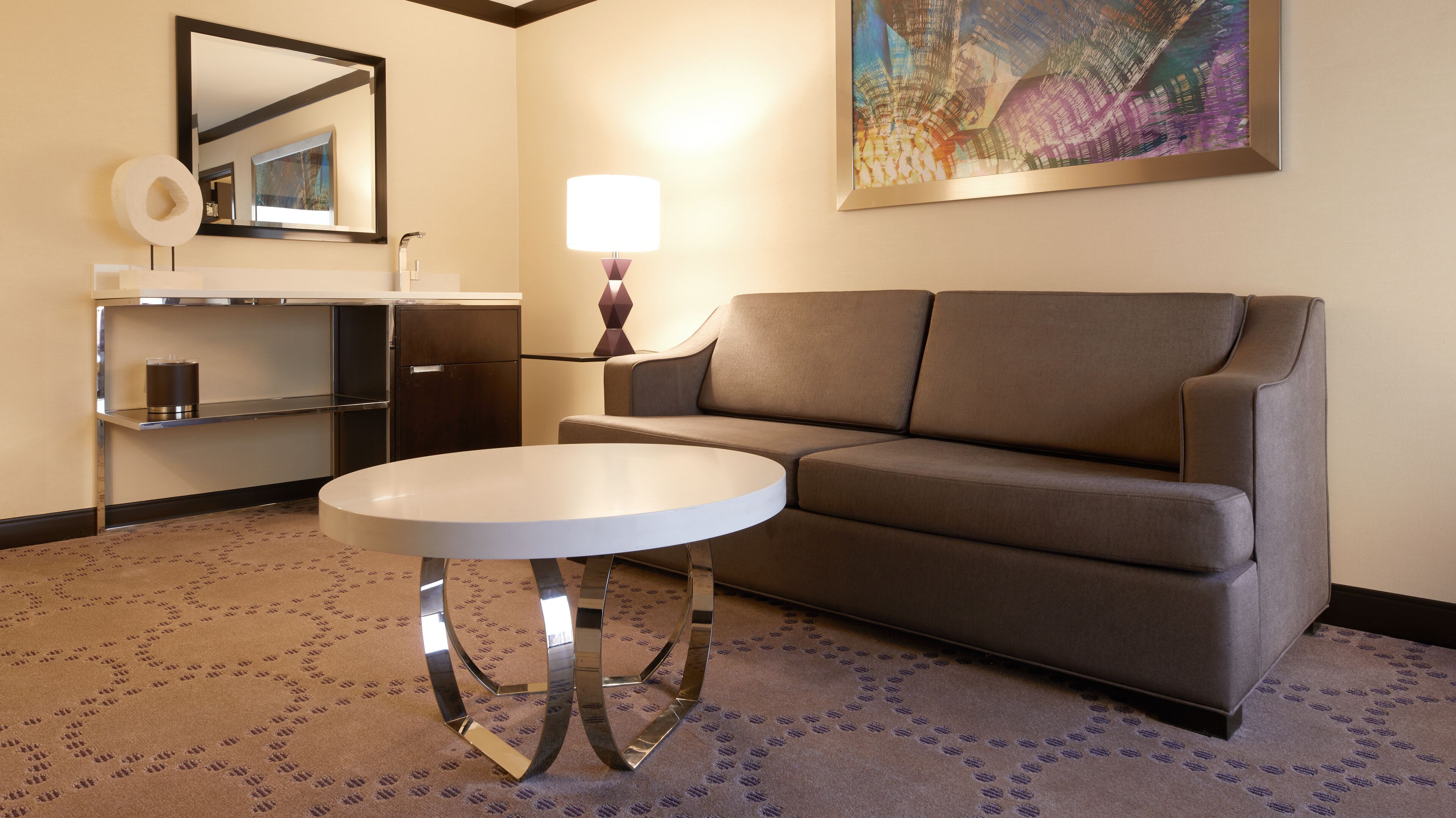 A modern hotel room with a sofa, a round table, a mirror, a lamp, and a painting on the wall.