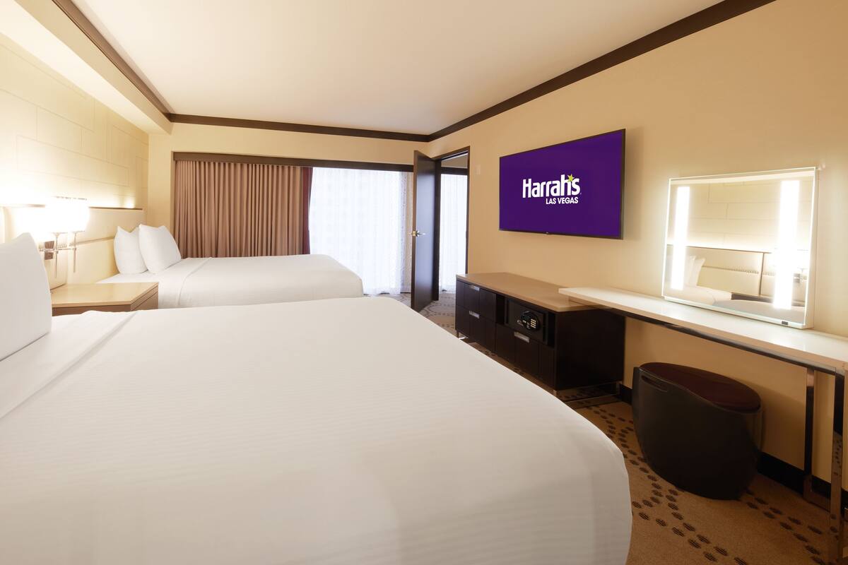 A hotel room with two beds, a desk, a chair, a trash bin, and a Harrah's Las Vegas sign.