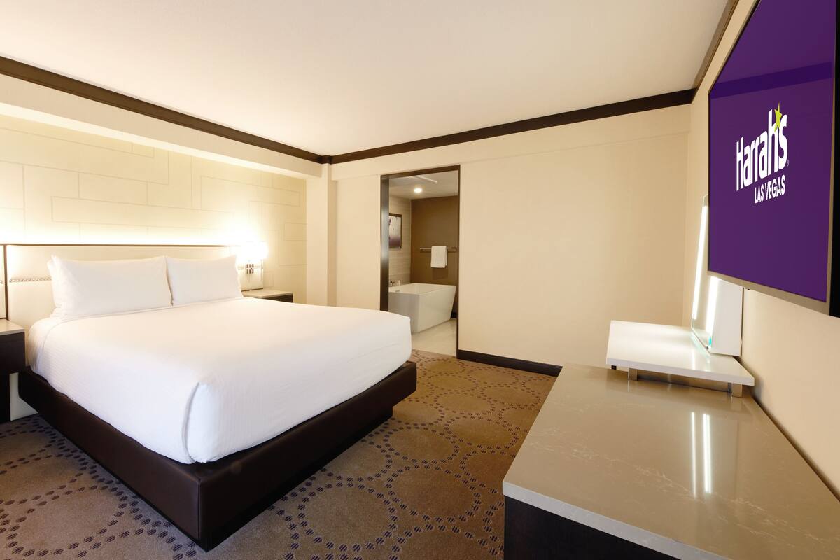 A hotel room with a large bed, a nightstand, a bathroom visible through an open door, and a Harrah's Las Vegas sign on the wall.