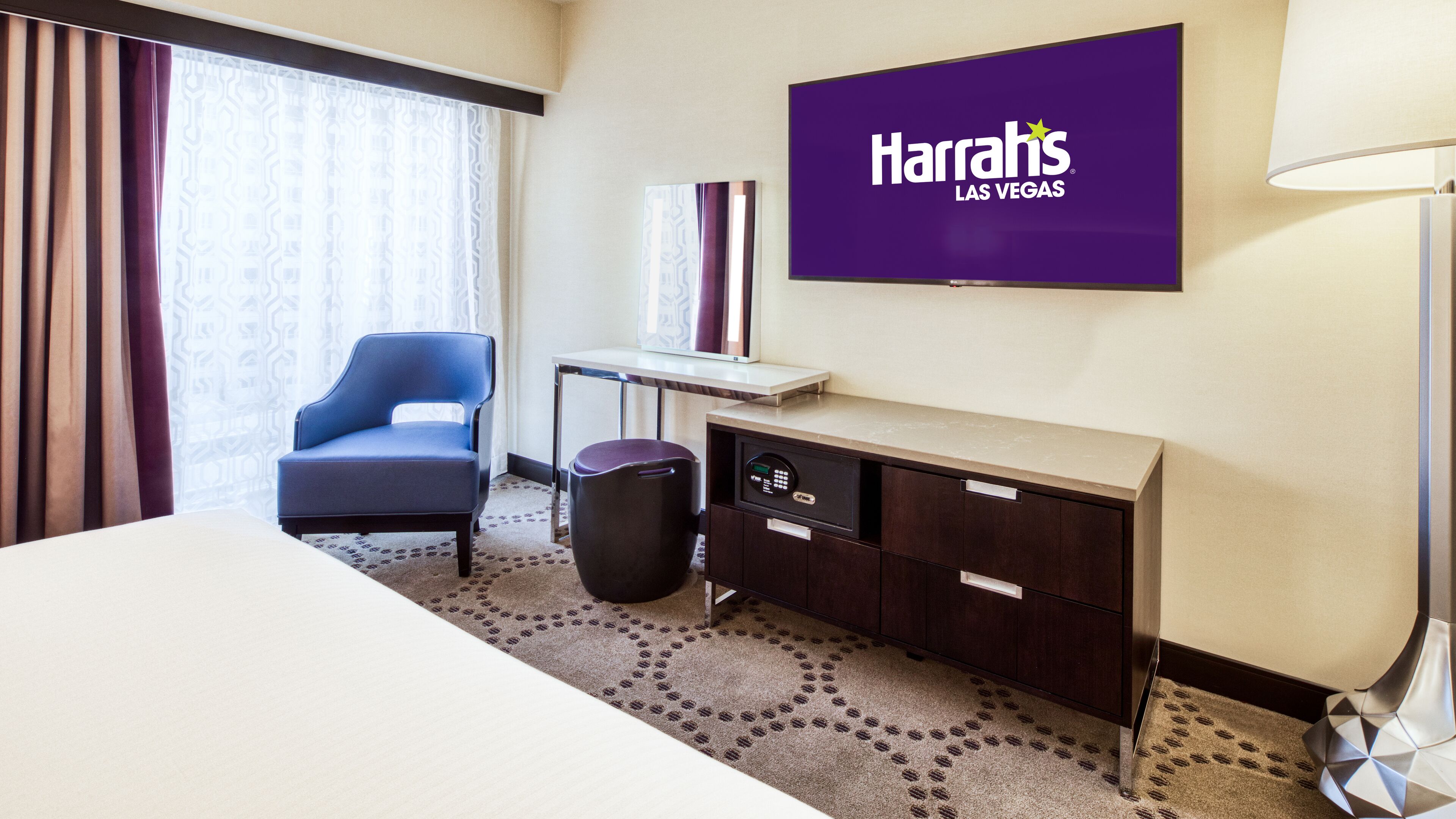 A hotel room with a bed, a desk, a chair, a trash bin, and a Harrah's Las Vegas sign on the wall.