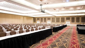 Un amplio salón de conferencias con filas de mesas y sillas, una alfombra estampada y un techo alto con iluminación empotrada.