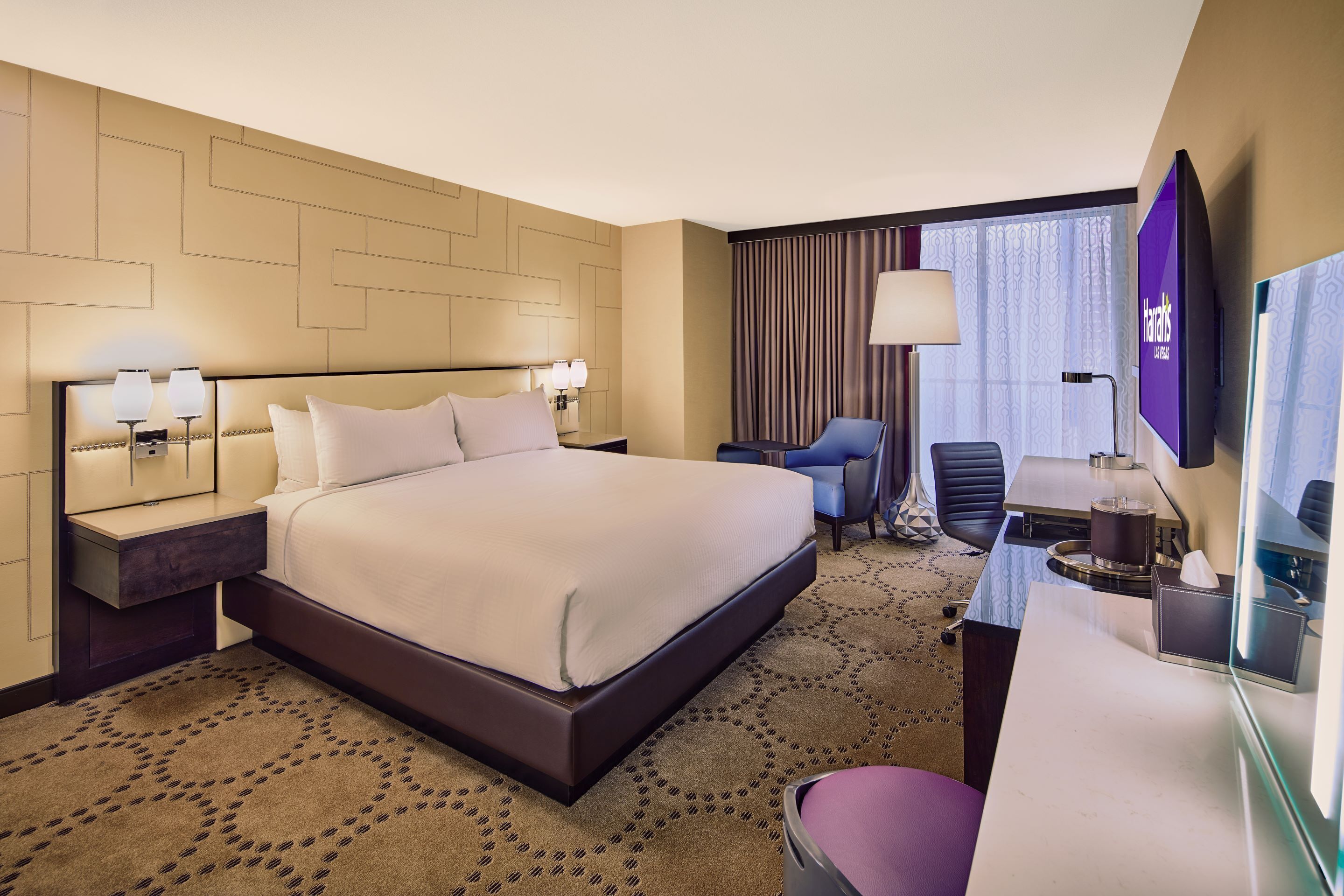 Ein Hotelzimmer mit einem großen Bett, einem Schreibtisch mit Stuhl, einem Fernseher und einem Fenster mit Vorhängen.