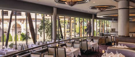 Un restaurant avec des tables dressées pour le service, de grandes fenêtres et une vue sur la ville.
