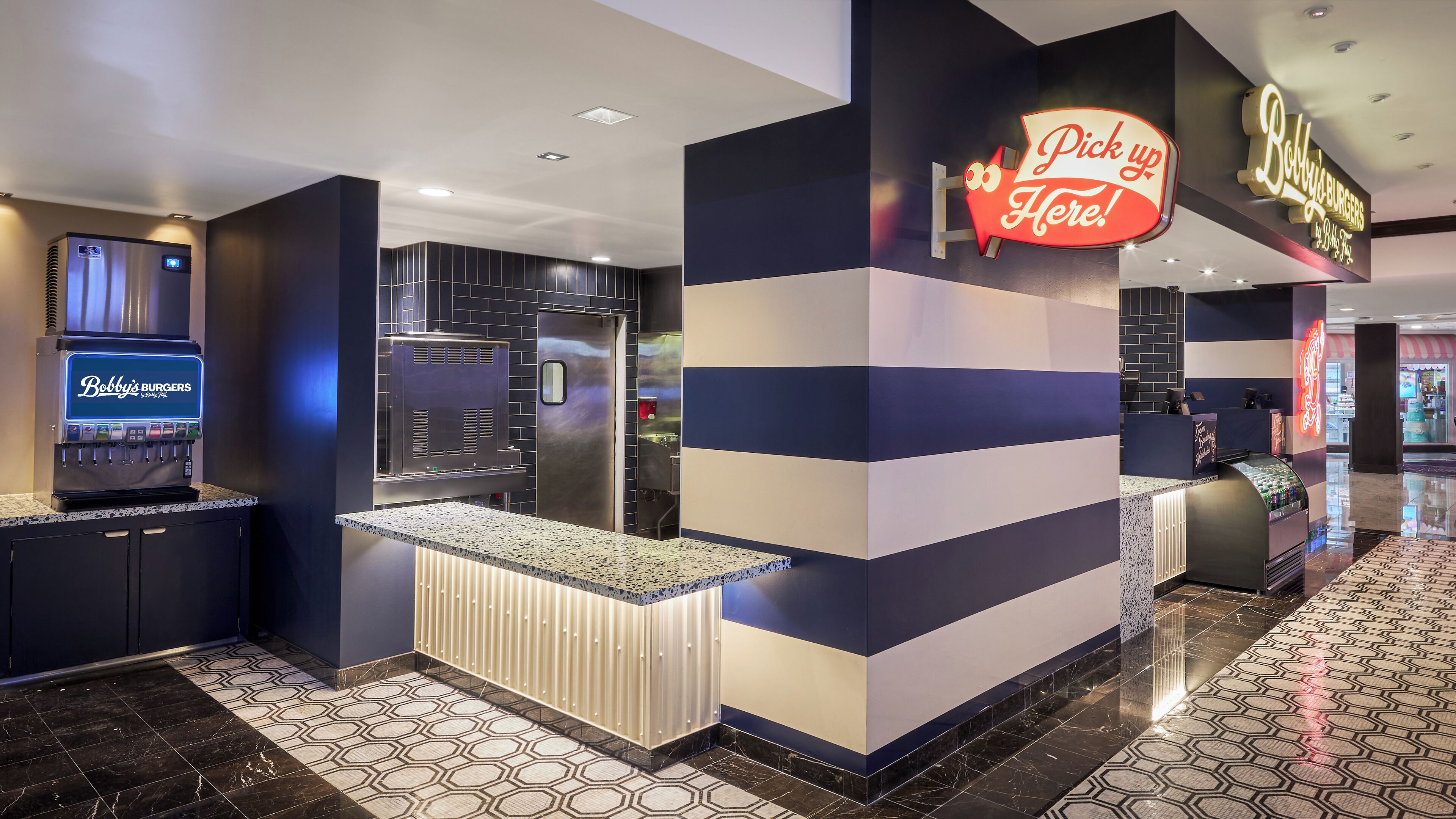 A modern kitchen area with a self-service beverage machine, a counter, and a neon sign indicating a pick-up point.