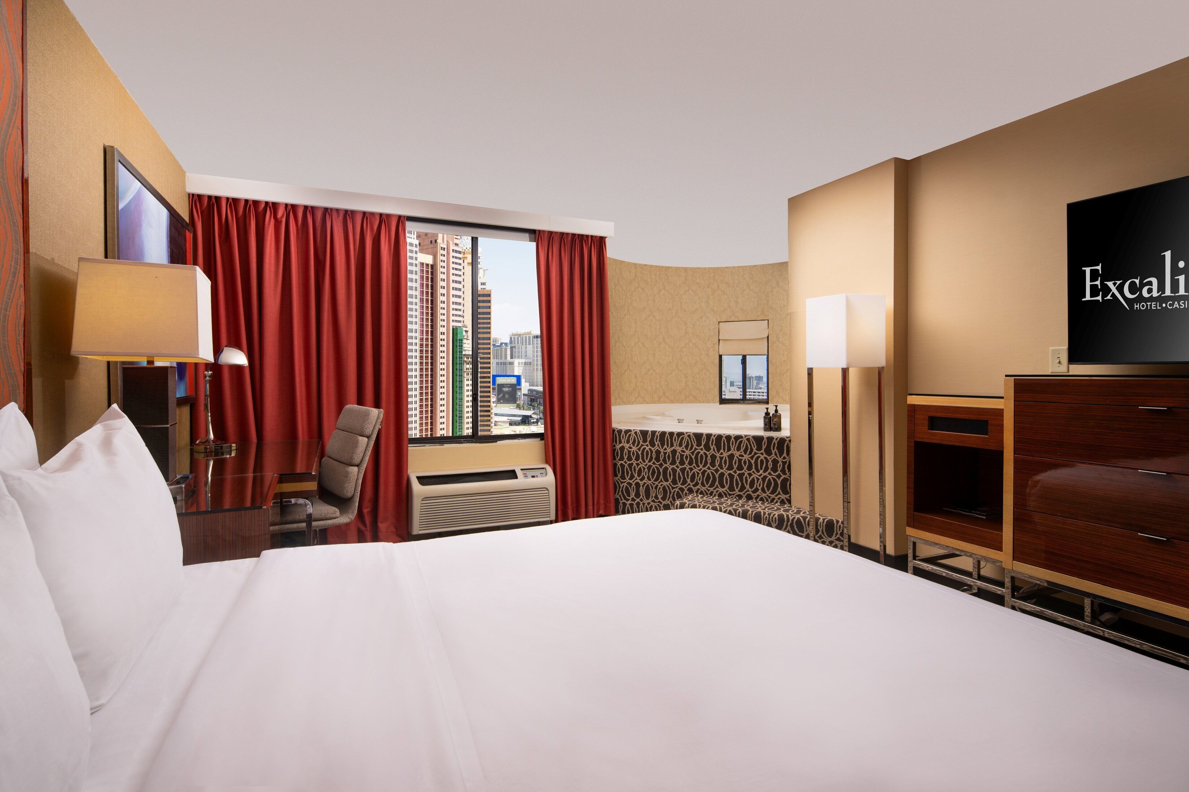 Habitación de hotel con cama, escritorio, silla, televisor, ventana con cortinas y vistas de la ciudad.