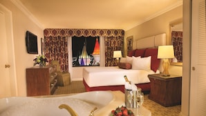A hotel room with a large bathtub, a bed with red pillows, a nightstand with a lamp, and a view of a lit tower at night.