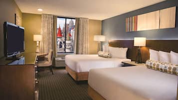 A hotel room with two beds, a desk, a TV, and a view of a cityscape.