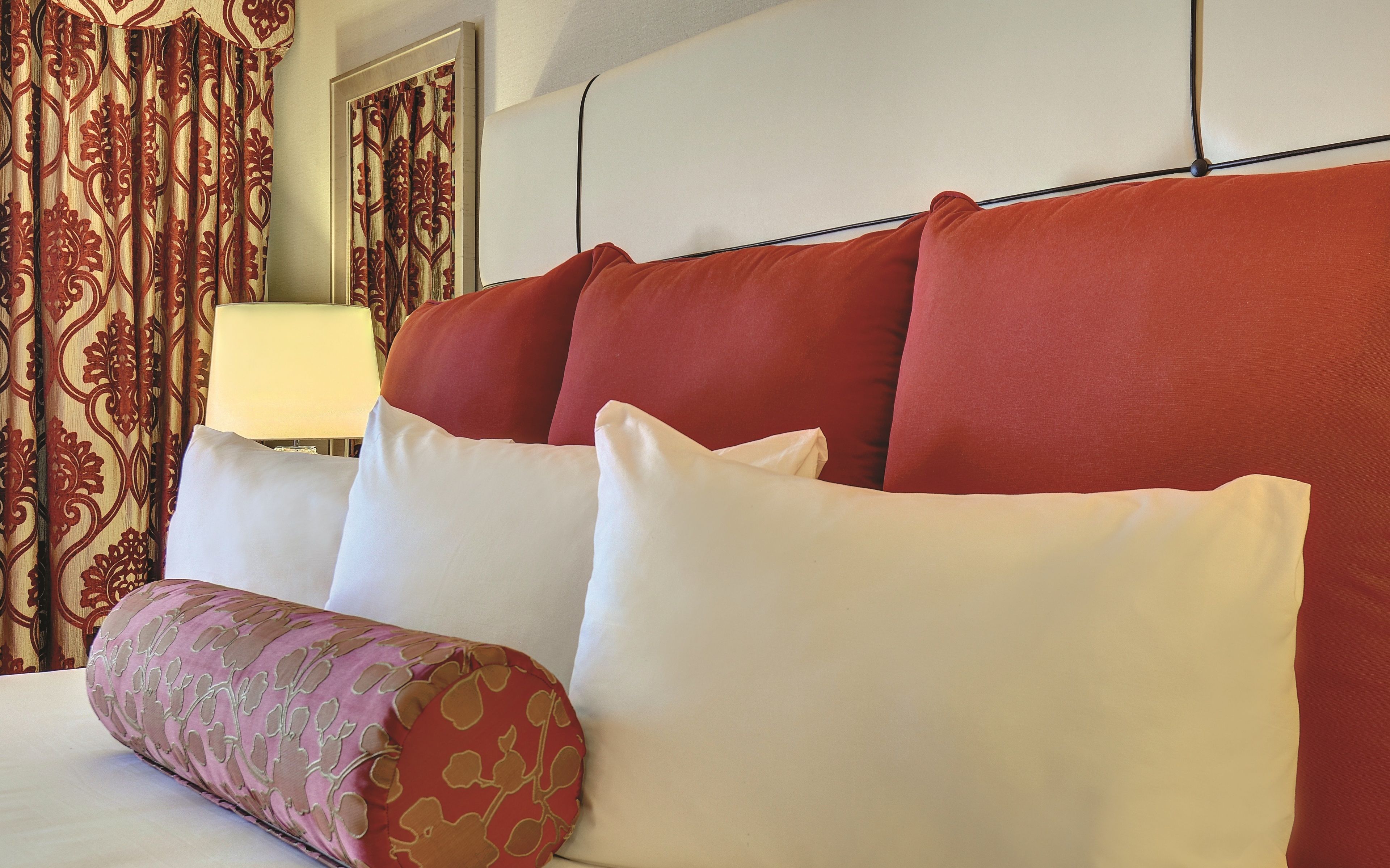 Habitación de hotel con una cama, almohadas rojas y blancas, un cojín decorativo estampado y una lámpara.
