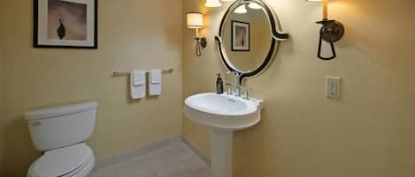 A bathroom with a toilet, sink, and mirror. There are wall-mounted lights and a framed picture on the wall.