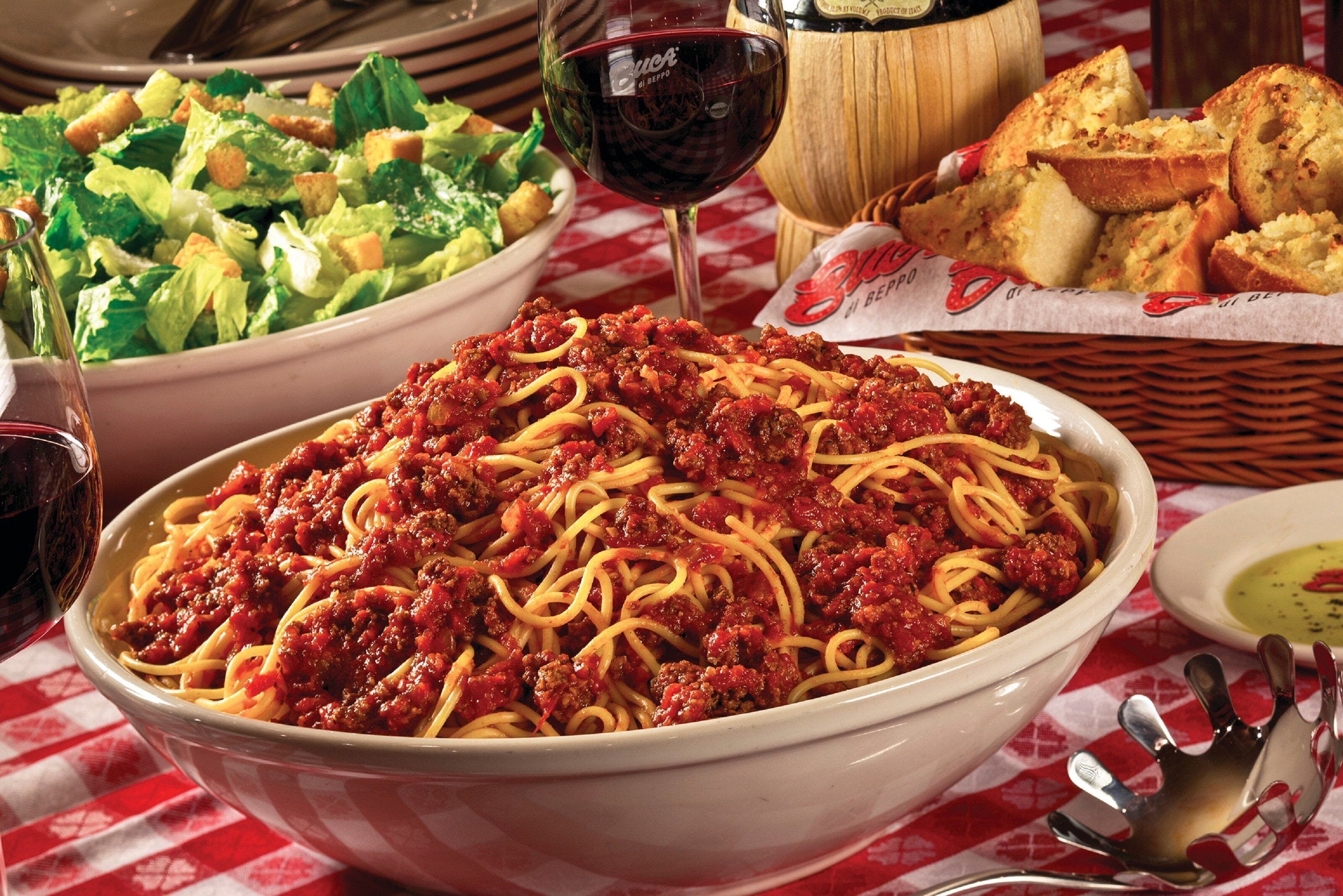 A bowl of spaghetti with meat sauce, a glass of red wine, a basket of bread, and a side salad on a table with a checkered tablecloth.