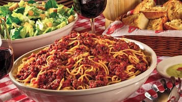 A bowl of spaghetti with meat sauce, a glass of red wine, a basket of bread, and a side salad on a table with a checkered tablecloth.
