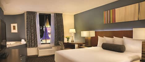 A hotel room with a large bed, a desk with a chair, a window with curtains, a wall-mounted TV, and a bedside table with a lamp.