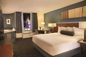 A hotel room with a large bed, a desk with a chair, a window with curtains, a wall-mounted TV, and a bedside table with a lamp. - Excalibur Hotel & Casino (Las Vegas)
