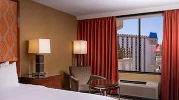 A hotel room with a bed, two bedside tables with lamps, a chair, a small round table, and a large window with red curtains.