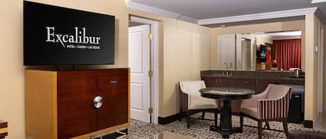 Ein Hotelzimmer mit Minibar, einem Fernseher, der 'Excalibur Hotel Casino Las Vegas' anzeigt, einer Essecke mit Tisch und Stühlen sowie einem gemusterten Teppich.