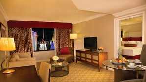 A hotel room with a sofa, a television, a dining table with fruit, and a view of a building outside.