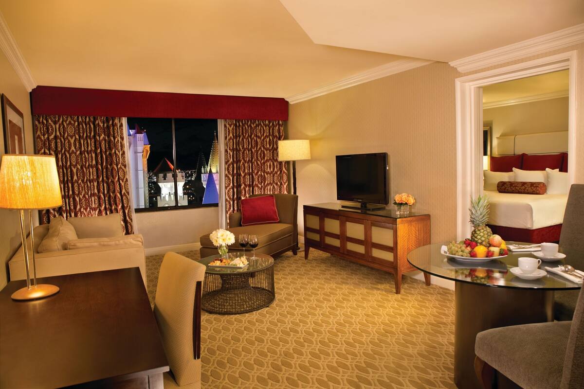 Een hotelkamer met een bank, een televisie, een eettafel met fruit en uitzicht op een gebouw.