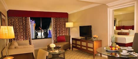 Een hotelkamer met een bank, een televisie, een eettafel met fruit en uitzicht op een gebouw.