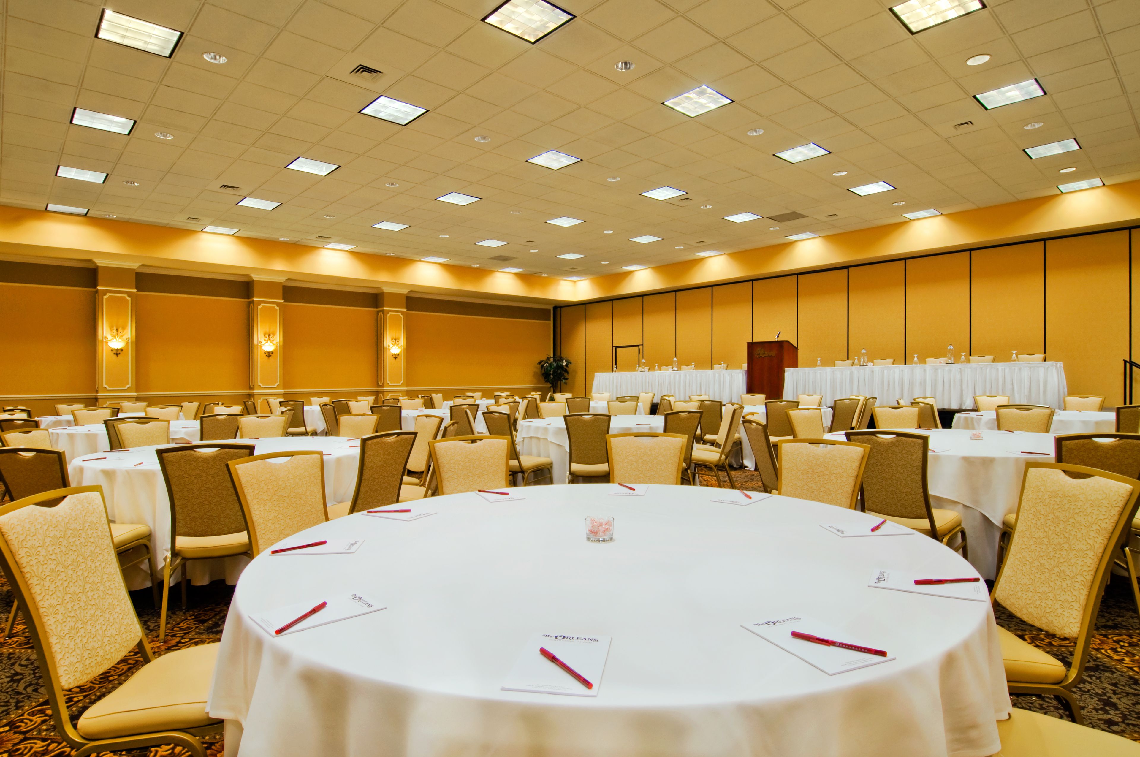 Een ruime congreszaal met meerdere ronde tafels, elk omringd door stoelen, en een podium.