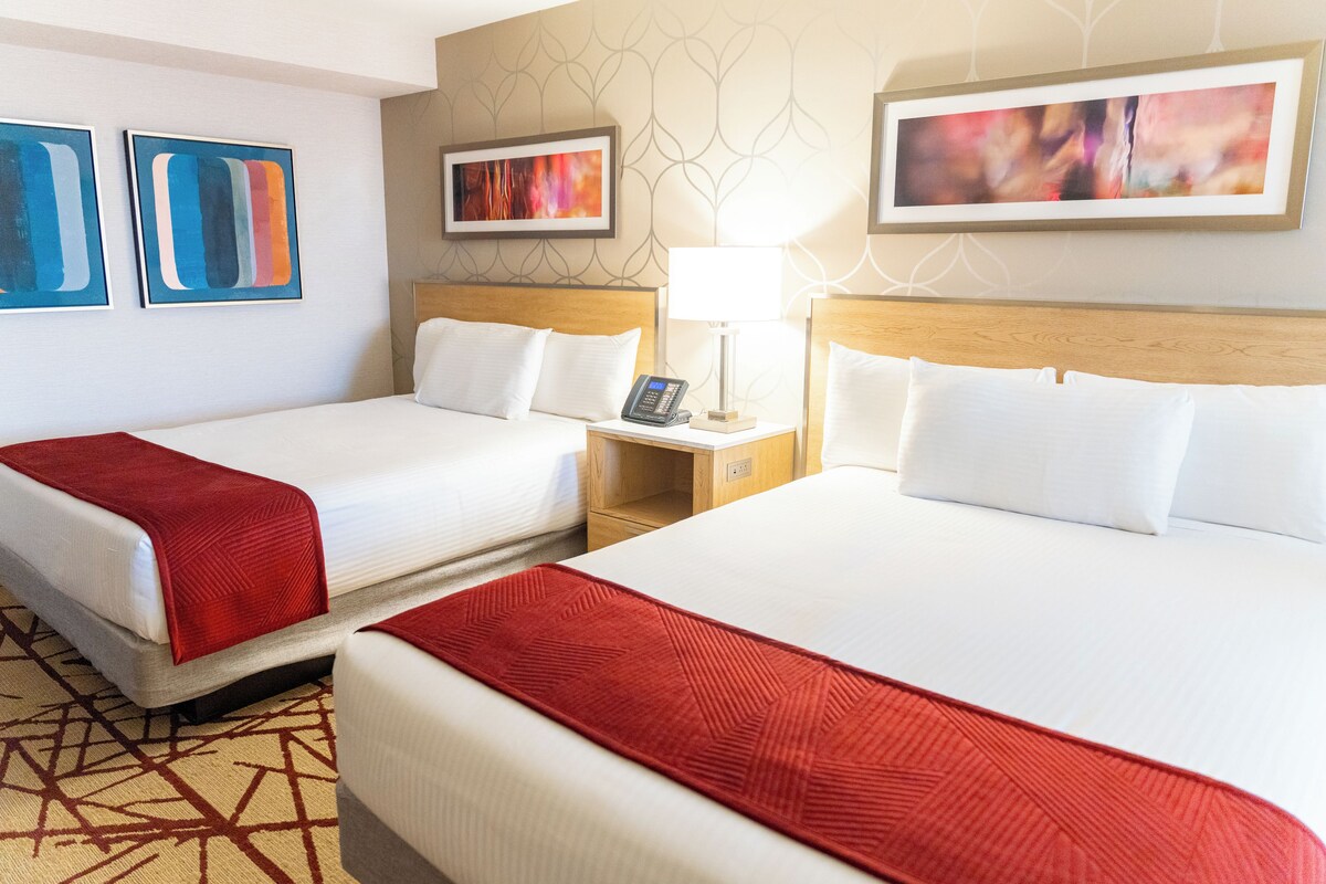 A hotel room with two beds, a nightstand with a lamp, and artwork on the walls.