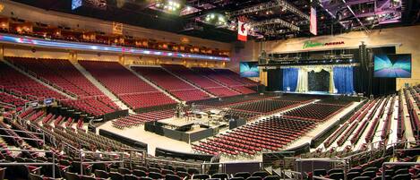 A large indoor auditorium with multiple rows of red seats, a stage at the front, and a large screen displaying an event.