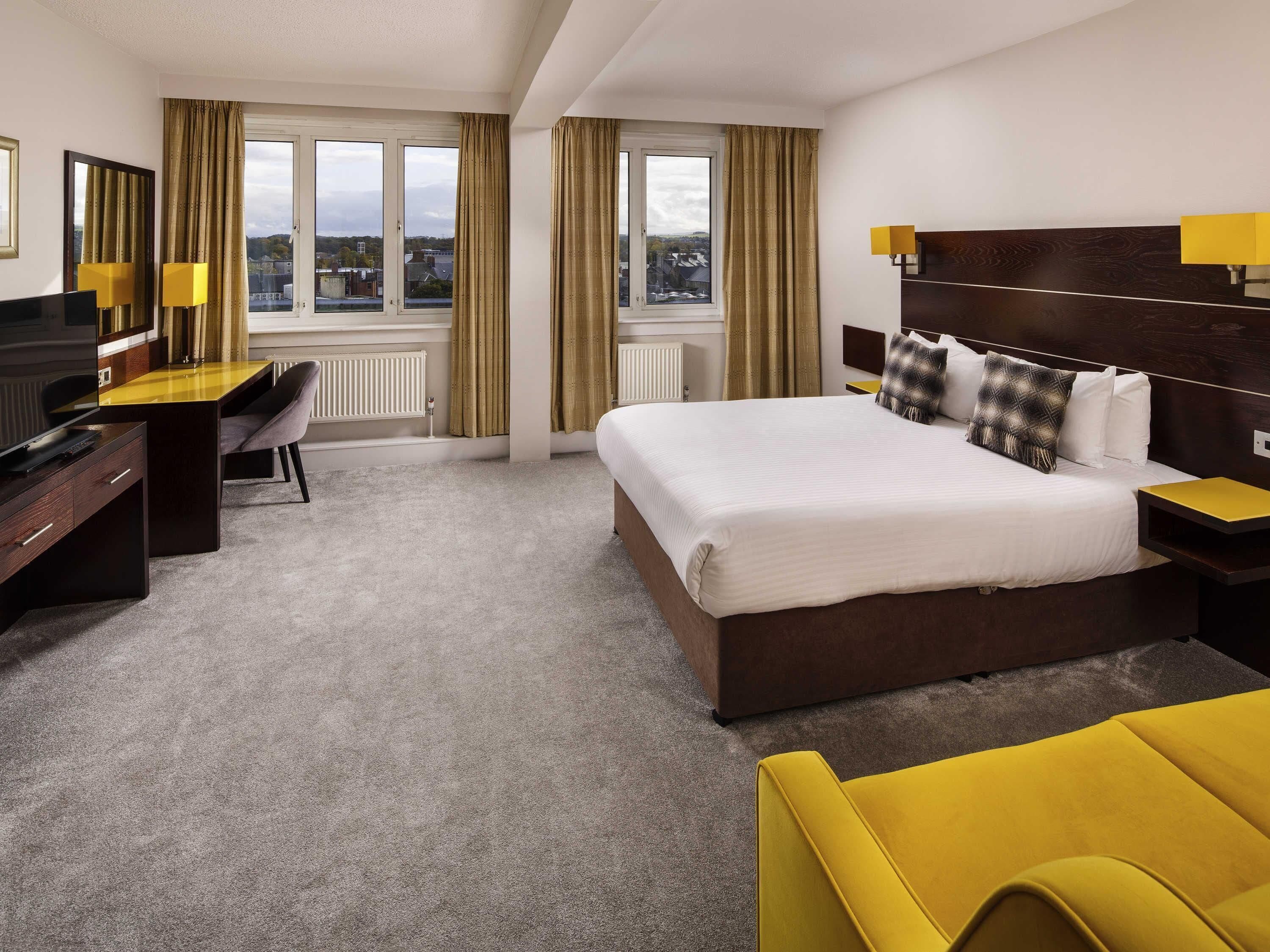 A hotel room with a large bed, a desk, a chair, a television, and a view of the city through the windows.
