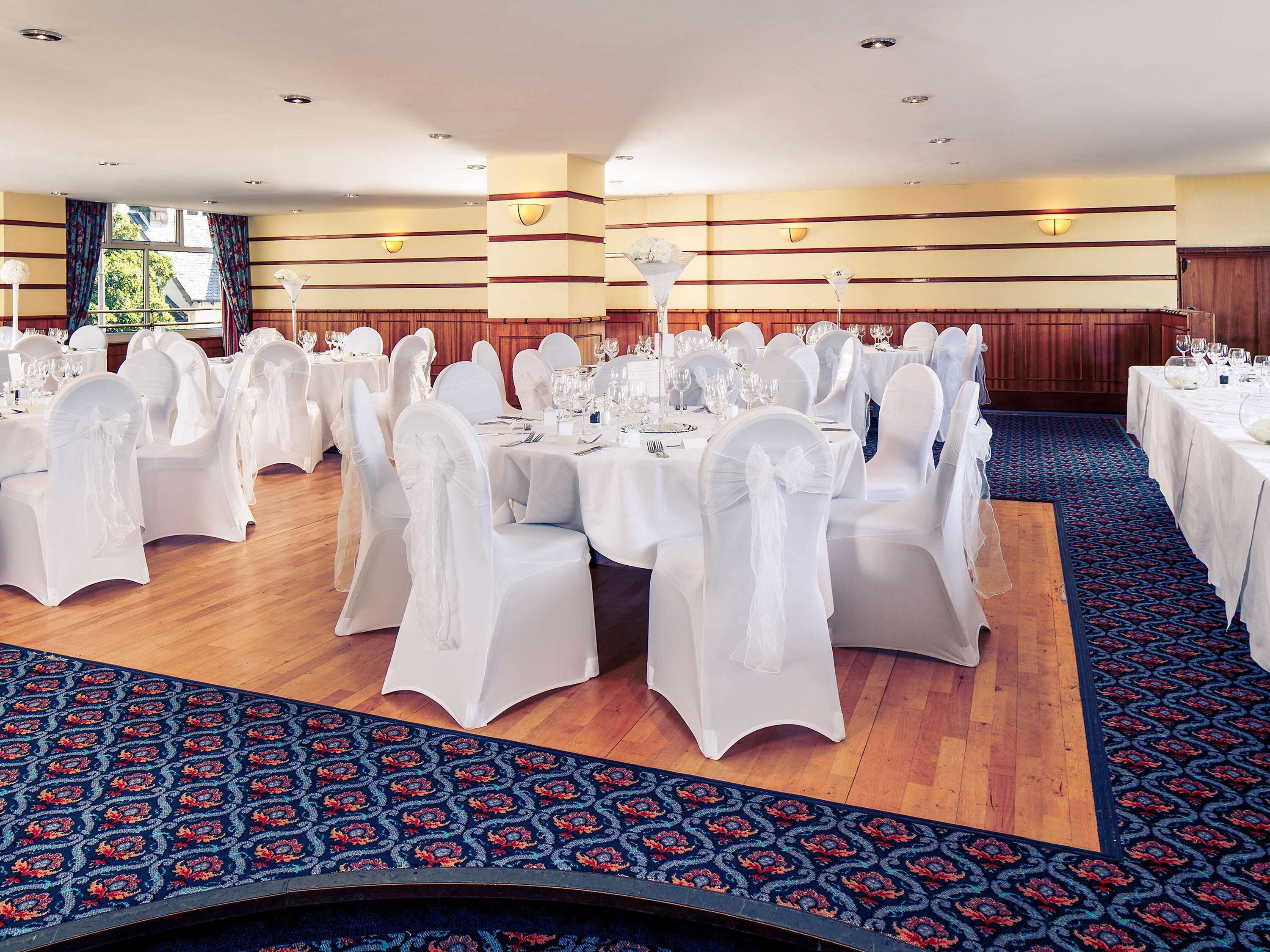 A banquet hall with round tables, white chairs, and a patterned carpet.