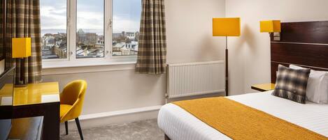 A hotel room with a bed, a desk, a chair, a window with curtains, a radiator, and two bedside lamps.