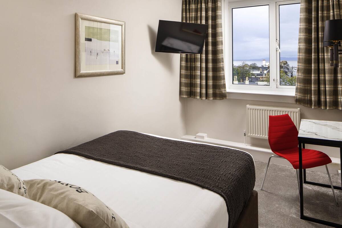 A hotel room with a bed, a desk, a chair, a TV, a window with curtains, and a radiator.