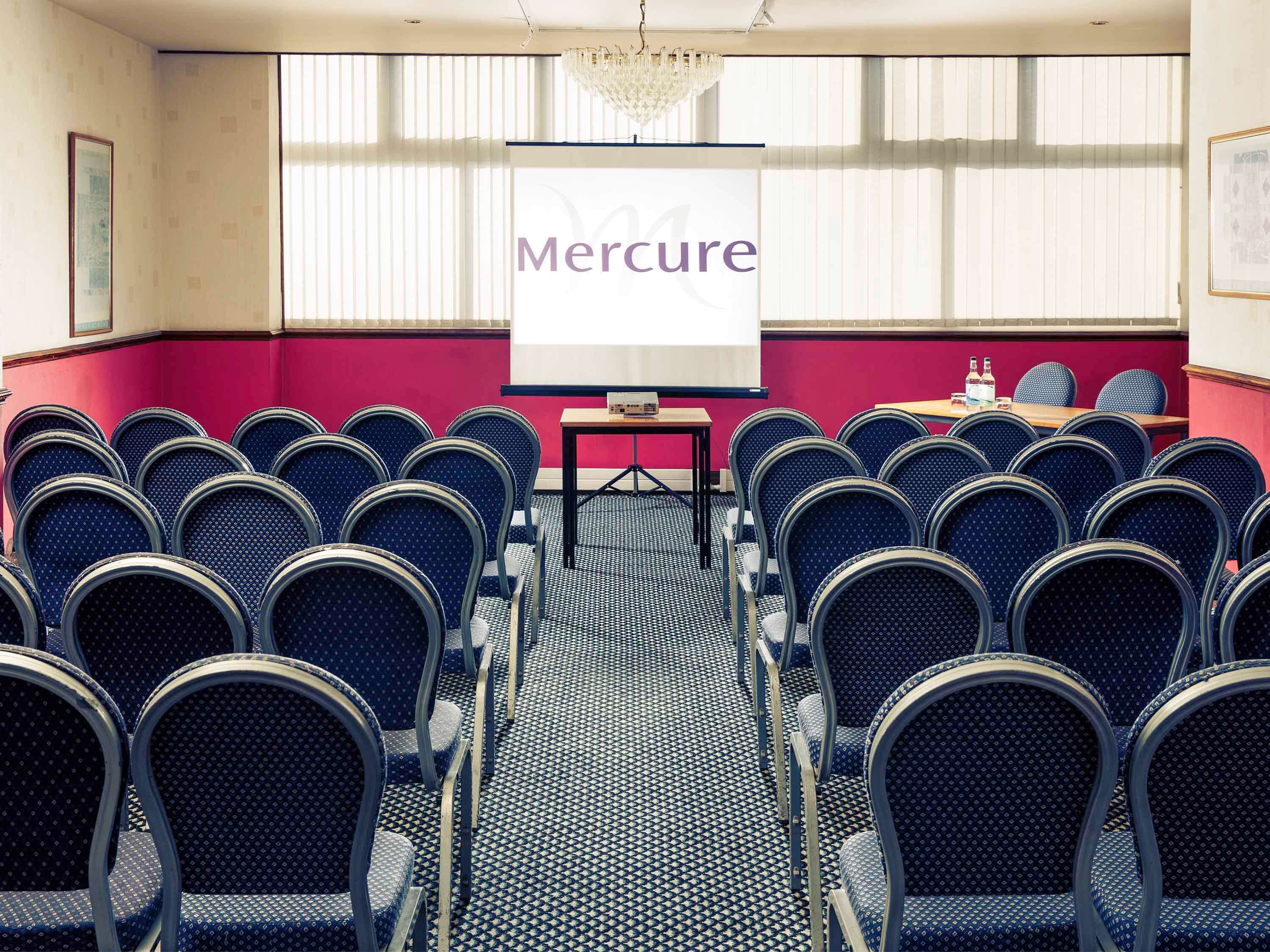 A conference room with rows of blue chairs, a projector screen with 'Mercure' on it, and a chandelier.