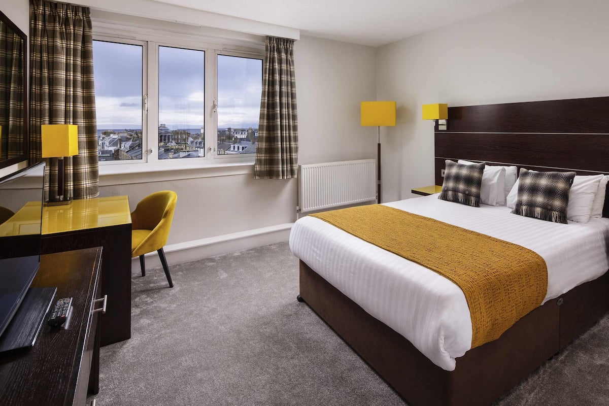 A hotel room with a bed, a desk, a chair, two lamps, a view through a window, and a radiator.