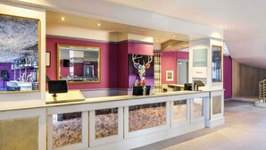 A reception area with a counter, a mirror, a colorful artwork, and a welcome sign. - Mercure Ayr Hotel (Ayr)