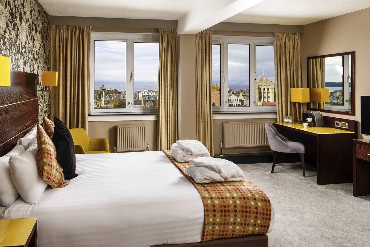 A hotel room with a bed, a desk, a chair, a radiator, and a view of the cityscape.