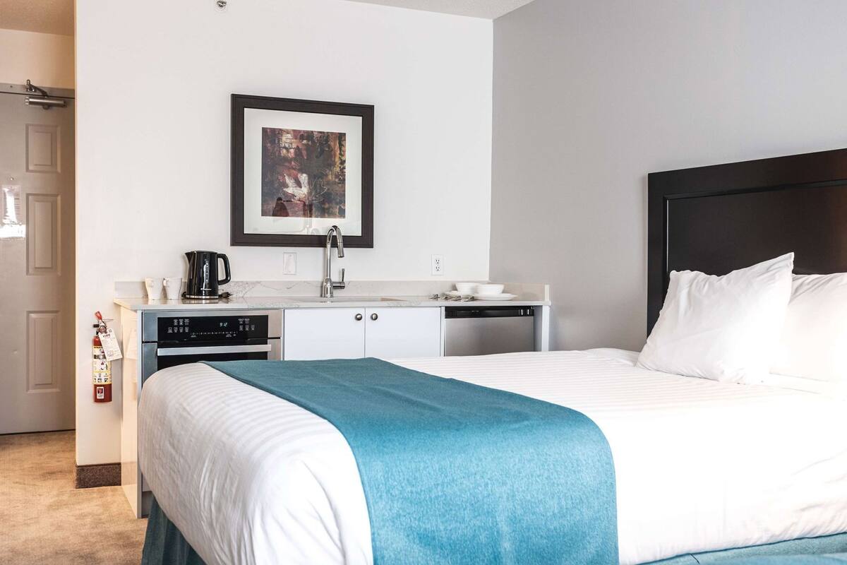 A hotel room with a bed, a small kitchenette, a framed picture on the wall, and a door.