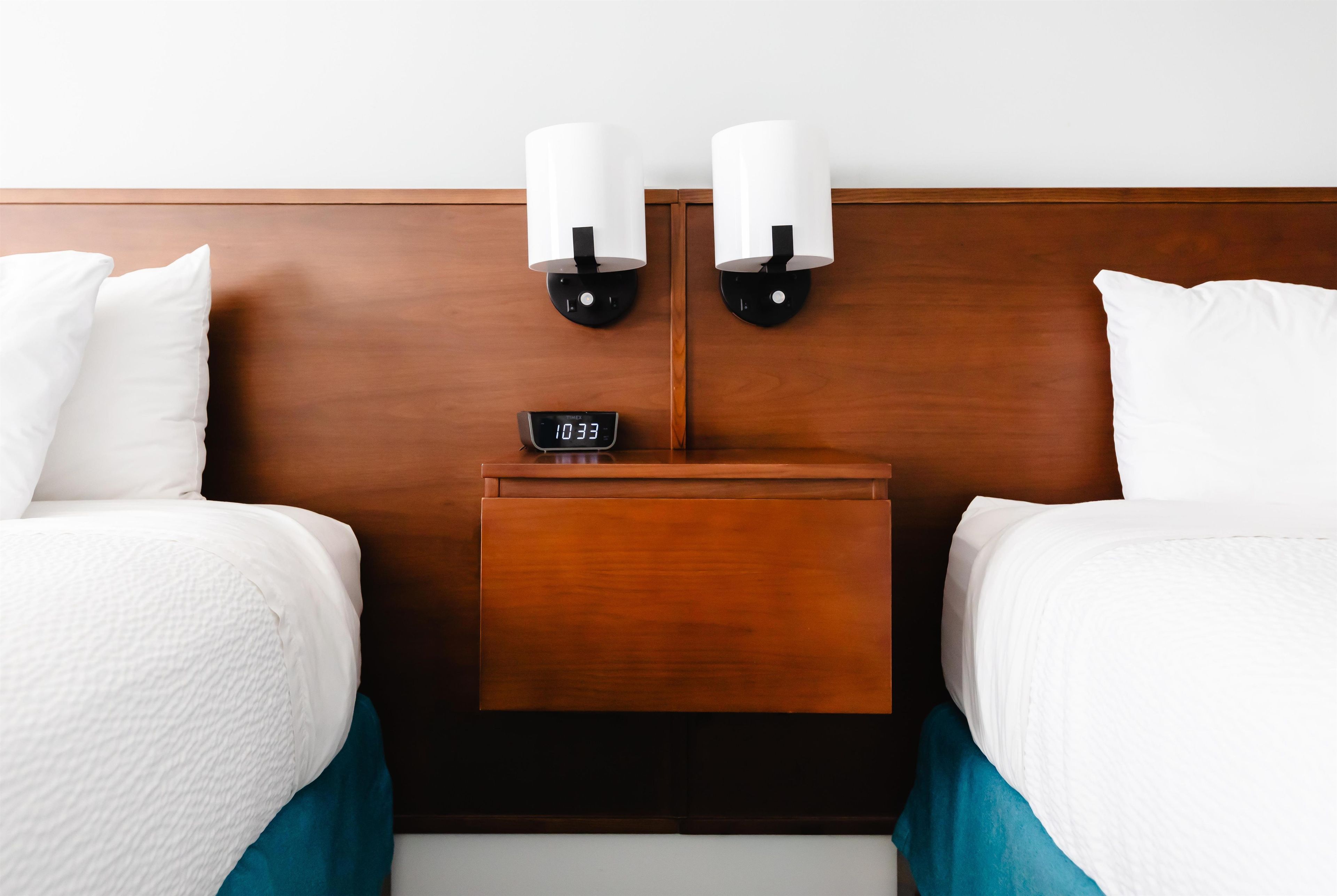 A hotel room with two beds, a nightstand with a digital clock, and wall-mounted lamps.