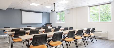 Un salón de conferencias con una gran mesa, sillas, una pantalla de proyector y ventanas.