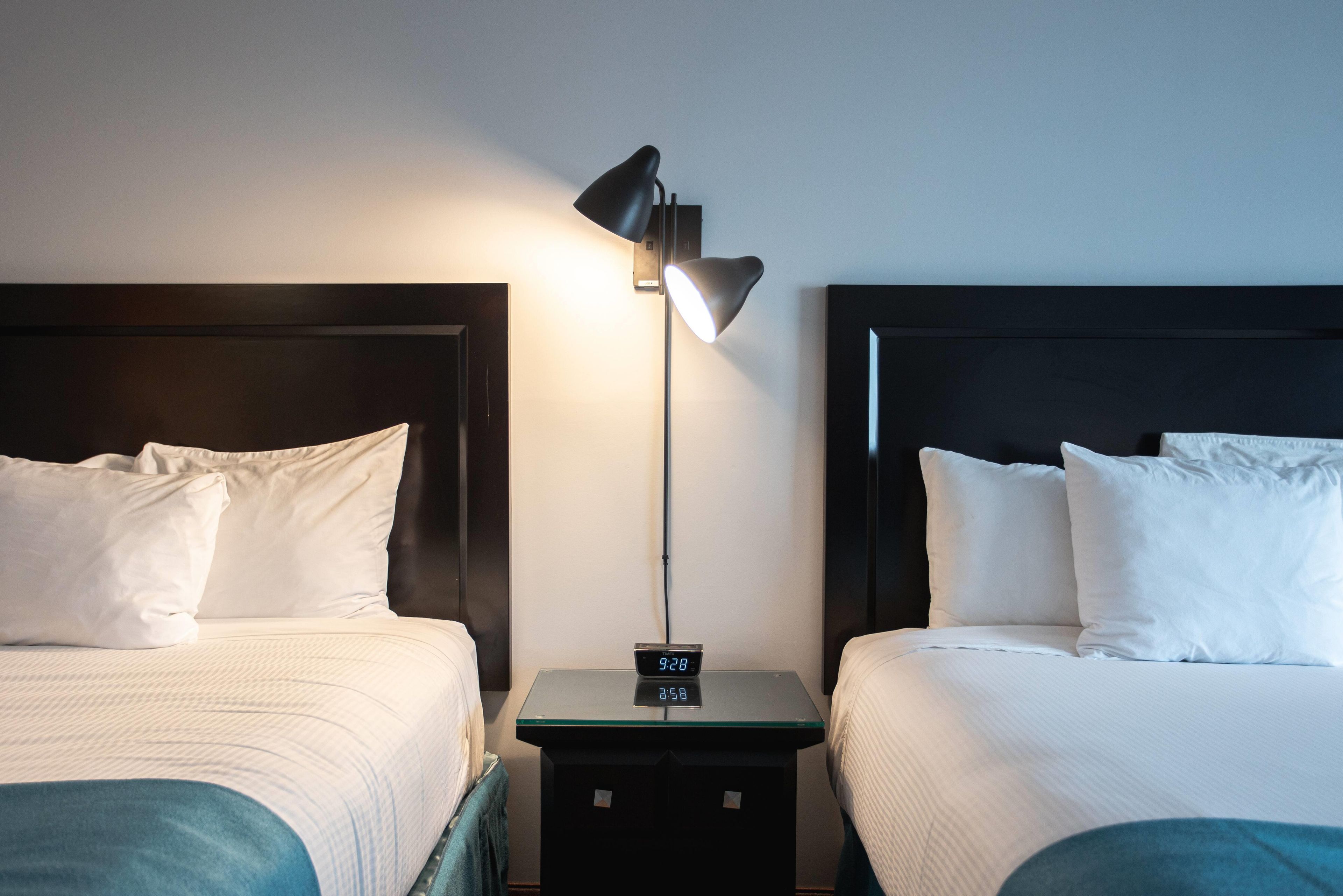 A hotel room with two beds, a nightstand with a digital clock, and a wall-mounted lamp.