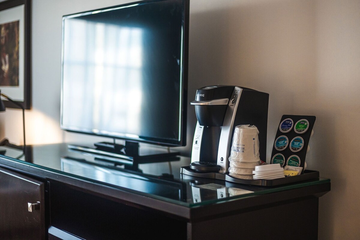 A hotel desk with a flat-screen TV, a coffee maker, a coffee pod holder, and a glass display case.