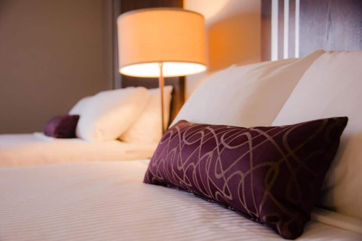 Una cama bien hecha con almohadas blancas y moradas, una lámpara en una mesita de noche y una pared con paneles de madera.