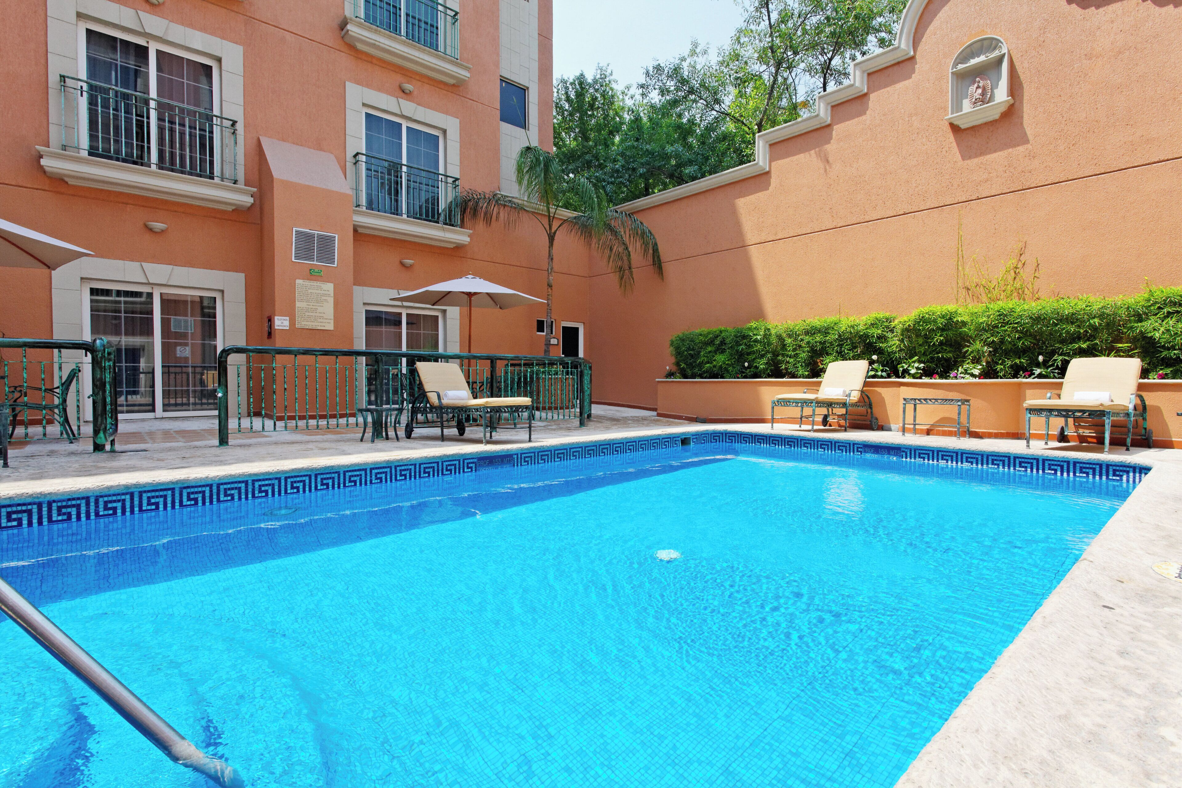 Une piscine entourée de chaises longues et agrémentée d’un parasol, le tout bordé d’un bâtiment doté de nombreuses fenêtres et de balcons.
