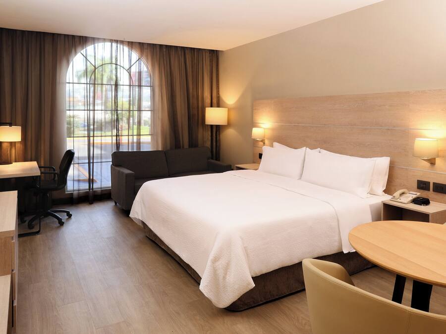 Een hotelkamer met een groot bed, een bureau met stoel, een klein tafeltje en een raam met gordijnen.