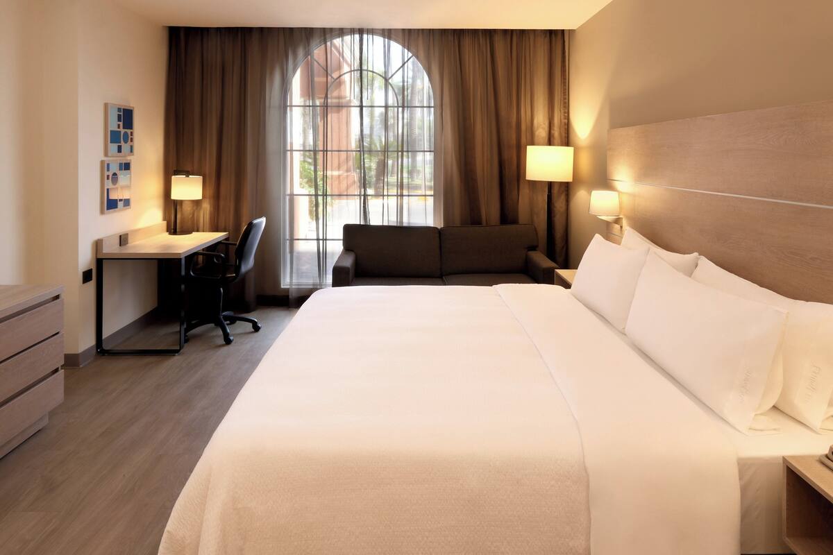 Una habitación de hotel con una cama grande, un escritorio, una silla y una ventana con cortinas.
